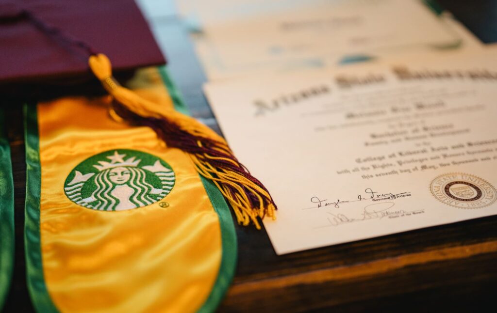 A burgundy graduation cap with a gold tassel and  Starbucks graduation stole next to a diploma 
