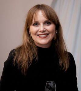 Caroline Caneva A woman with straight, light brown hair and bangs is smiling, wearing a black top and gold earrings, holding a glass with a light-colored drink, against a neutral background with soft lighting.