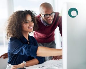Two colleagues smiling and looking at a computer screen together in an office setting, with one pointing at the monitor. A teal logo is visible on the wall in the background.