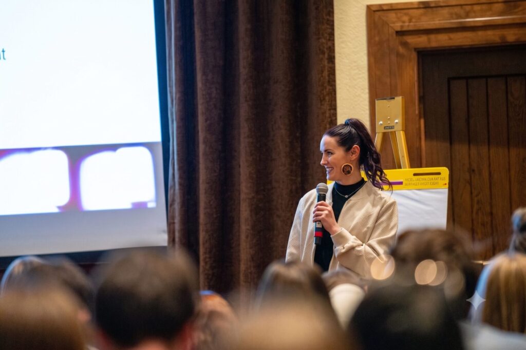 Rachelle Ray holds a microphone as she speaks to an audience in a conference room, standing near a projection screen and a yellow easel pad. The crowd listens attentively.