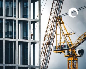 A yellow construction crane stands beside a modern concrete building with a grid-like facade, symbolizing progress and procurement reform. A worker is visible on the crane, and a circular logo appears in the top right corner against a cloudy sky.