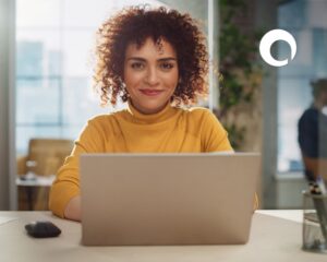 A woman with curly hair, wearing a yellow turtleneck, sits at a desk and smiles at the camera while using a laptop to learn about mistakes to avoid in proposal writing. A computer mouse and pen holder are on the desk in a bright office with a large window.