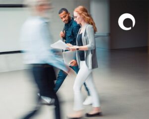 Two people walk together indoors, one holding papers, the other a laptop—clearly focused on strategies to drive revenue. Both appear in motion blur, with another blurred figure walking past. A white, circular logo is visible in the top right corner.
