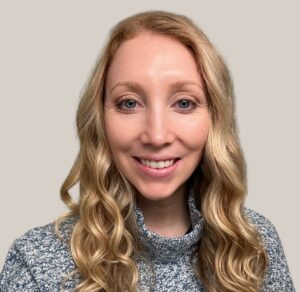 Janelle Poisel, a woman with long, wavy blonde hair, smiles at the camera. She is wearing a blue-gray, high-neck sweater and is pictured against a plain, light-colored background.