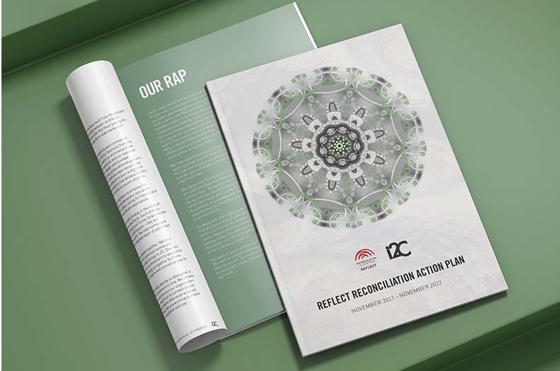 Two magazines lie on a green surface. The cover shows a decorative circular design and the title “Reflect RAP Reconciliation Action Plan November 2021 – November 2022” along with two logos at the bottom.