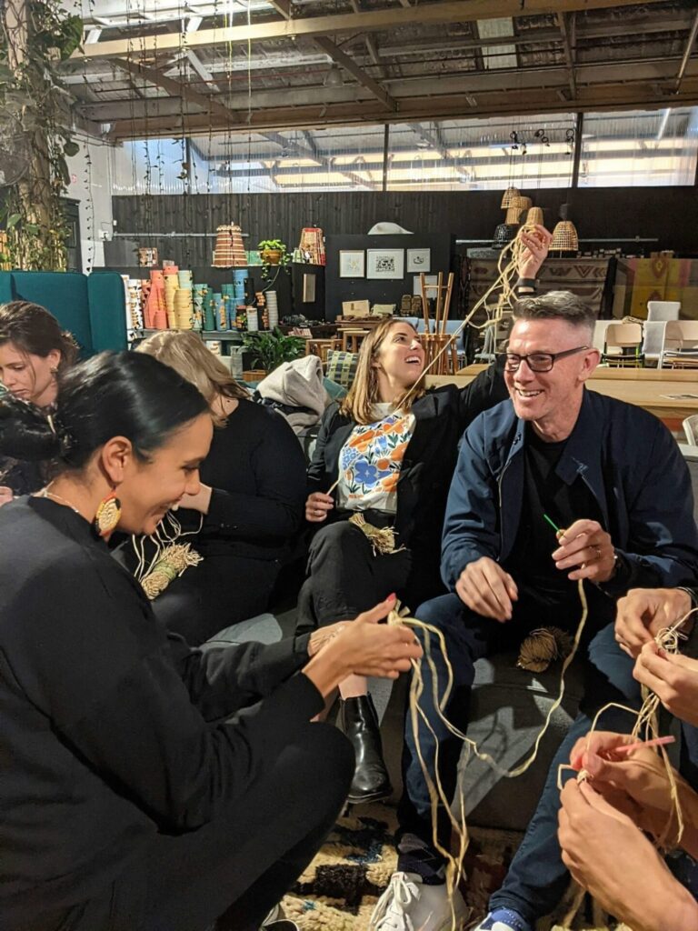 A group of people sit closely together indoors, laughing and weaving string as upbeat RAP music plays in the background. Yarn and craft supplies are visible nearby, adding to the lively and joyful atmosphere.