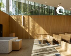 Sunlight streams through large windows onto wooden benches in a modern, minimalist room with light wood walls and floors. Greenery hangs outside, and geometric shadows fall inside—a space worthy of INDE.Awards recognition for its refined design.