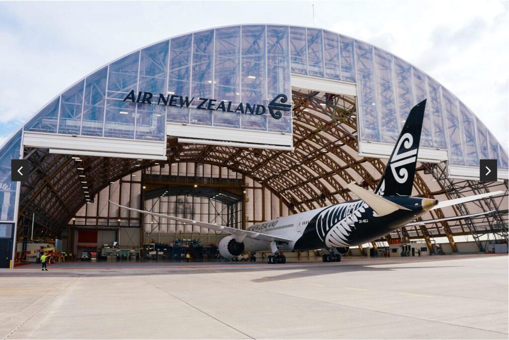 A black-and-white Air New Zealand airplane with a fern design is parked outside a large open hangar that has Air New Zealand written on it. The sky is partly cloudy.
