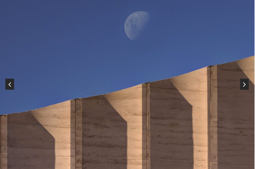 A waxing gibbous moon rises in a clear blue sky above a sunlit, wavy-edged concrete wall casting sharp shadows.
