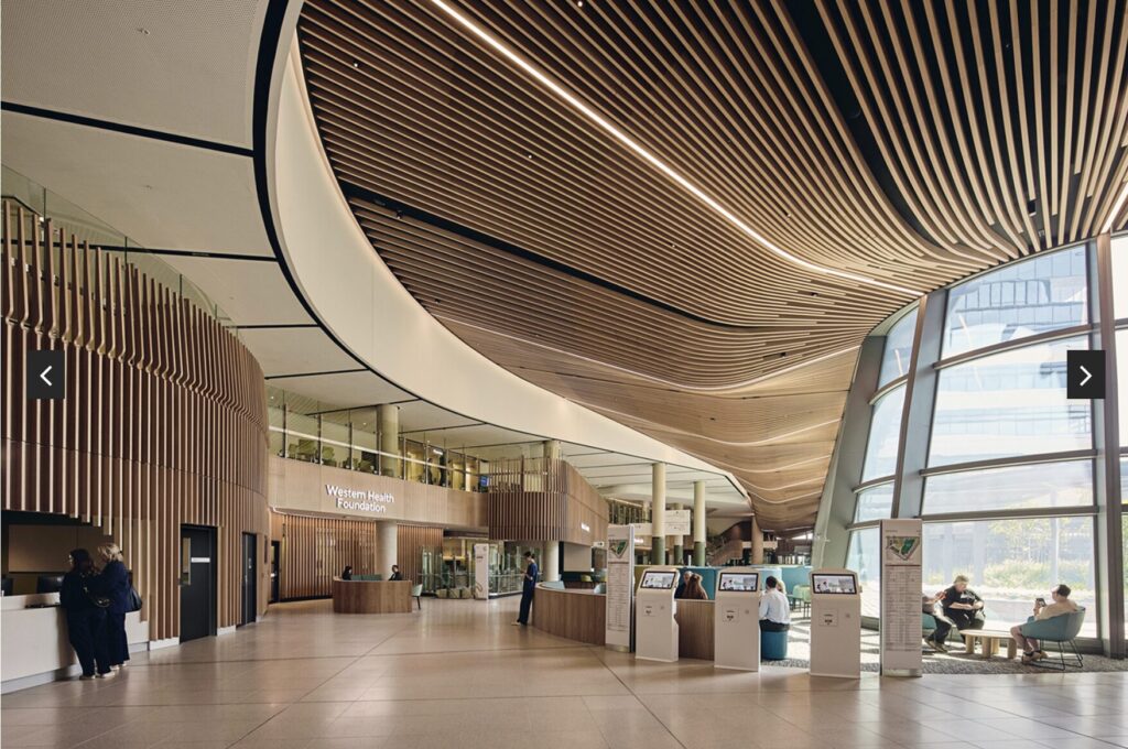 Spacious modern lobby with wood slat ceilings, curved architecture, large windows, information desks, self-service kiosks, and people sitting and walking. Sign reads Western Health Foundation.
