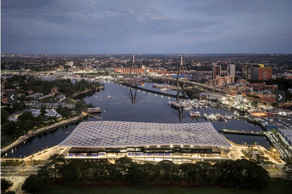 Aerial view of a modern, angular-roofed building by a river at dusk, with bridges, boats, and city buildings in the background under a cloudy sky.