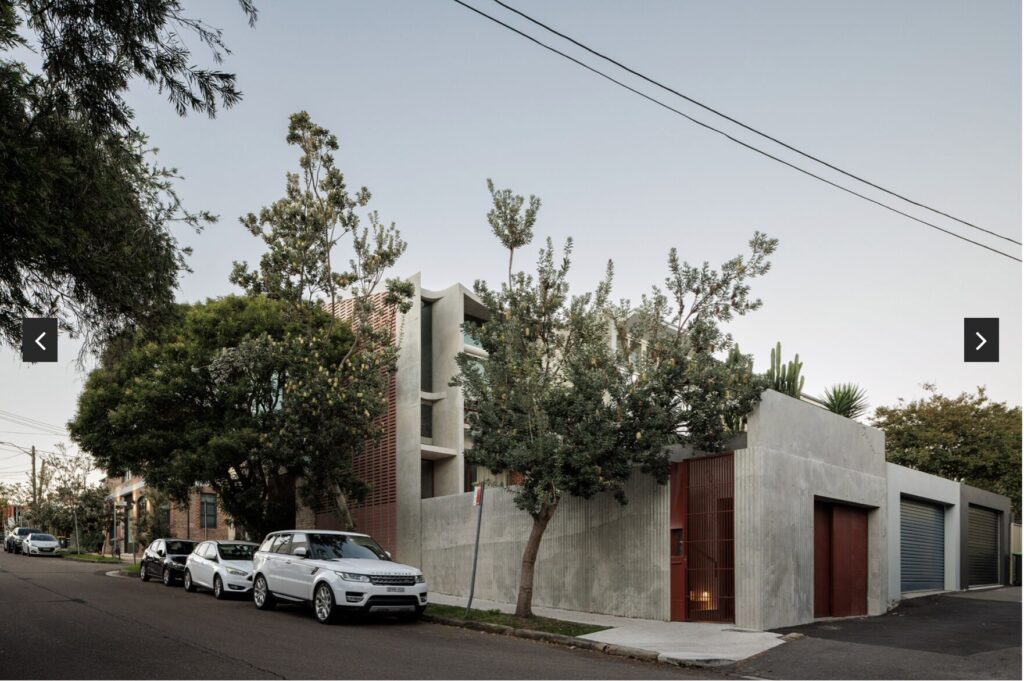 A modern concrete house with a red door and garage, surrounded by trees, is on a quiet street with several parked cars and overhead power lines.