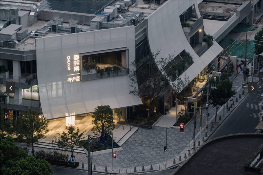 Modern building with curved white panels, large windows, and illuminated signage, situated on a street corner with trees and traffic lights. The surrounding area appears quiet with minimal pedestrian activity.