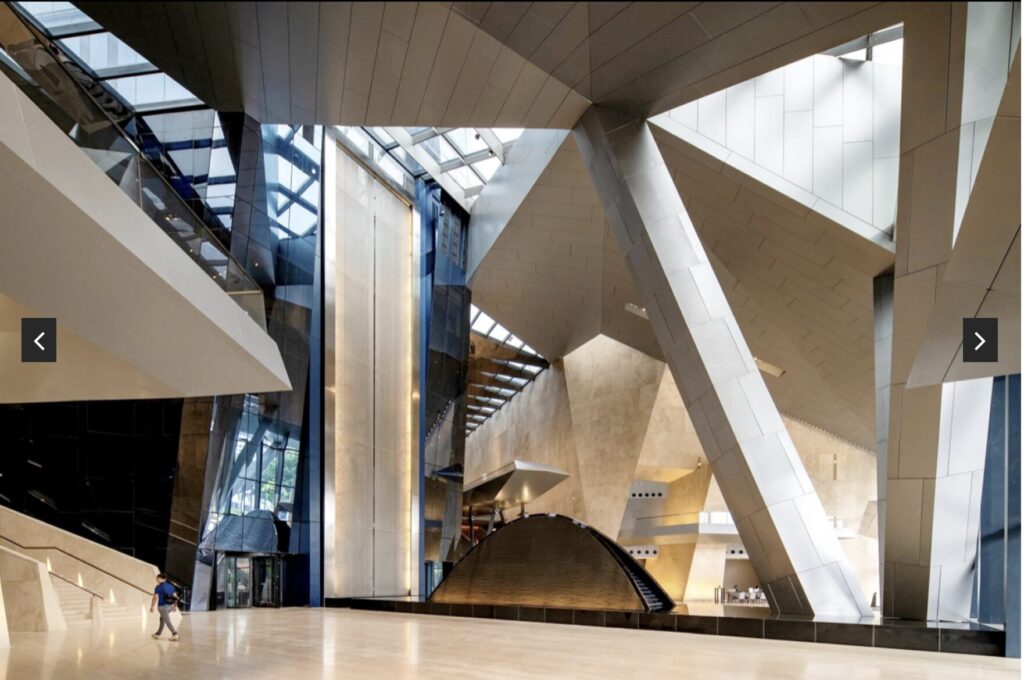 Spacious, modern interior with large angular beams, glass walls, and natural light. A person walks on the left side of the open floor, and stairs lead to upper levels. Architectural features create a futuristic look.