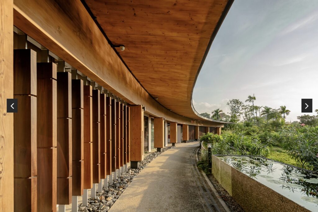 Curved walkway with a wooden roof, vertical wooden slats on the left, rocks along the edge, lush greenery and a reflecting pool on the right, under a partly cloudy sky.