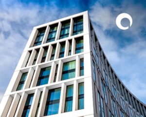 A modern, white multi-story building with large windows stands against a partly cloudy blue sky, reflecting sleek design worthy of architecture awards. A white circular logo appears in the top right corner of the image.