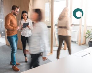A bustling office with two people walking past each other, blurred, in the foreground. In focus in the background, a man and a woman consult an ipad, looking focused