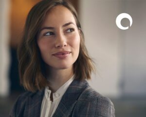 A woman with shoulder-length brown hair wearing a plaid blazer and white blouse looks to the side, smiling softly. A white circular logo, reminiscent of the OpenAsset vs. Bynder branding, is visible in the upper right corner against a blurred background.