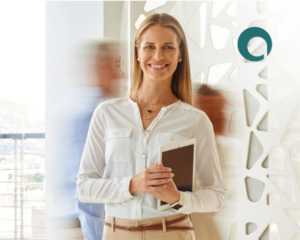 A blonde woman in an office is smiling and holding a notebook.